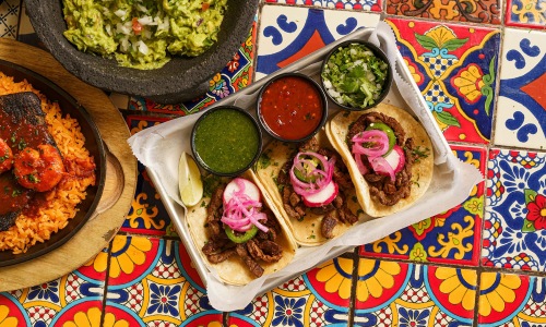 A tray of three tacos with salsa, guacamole, and rice on a colorful table.